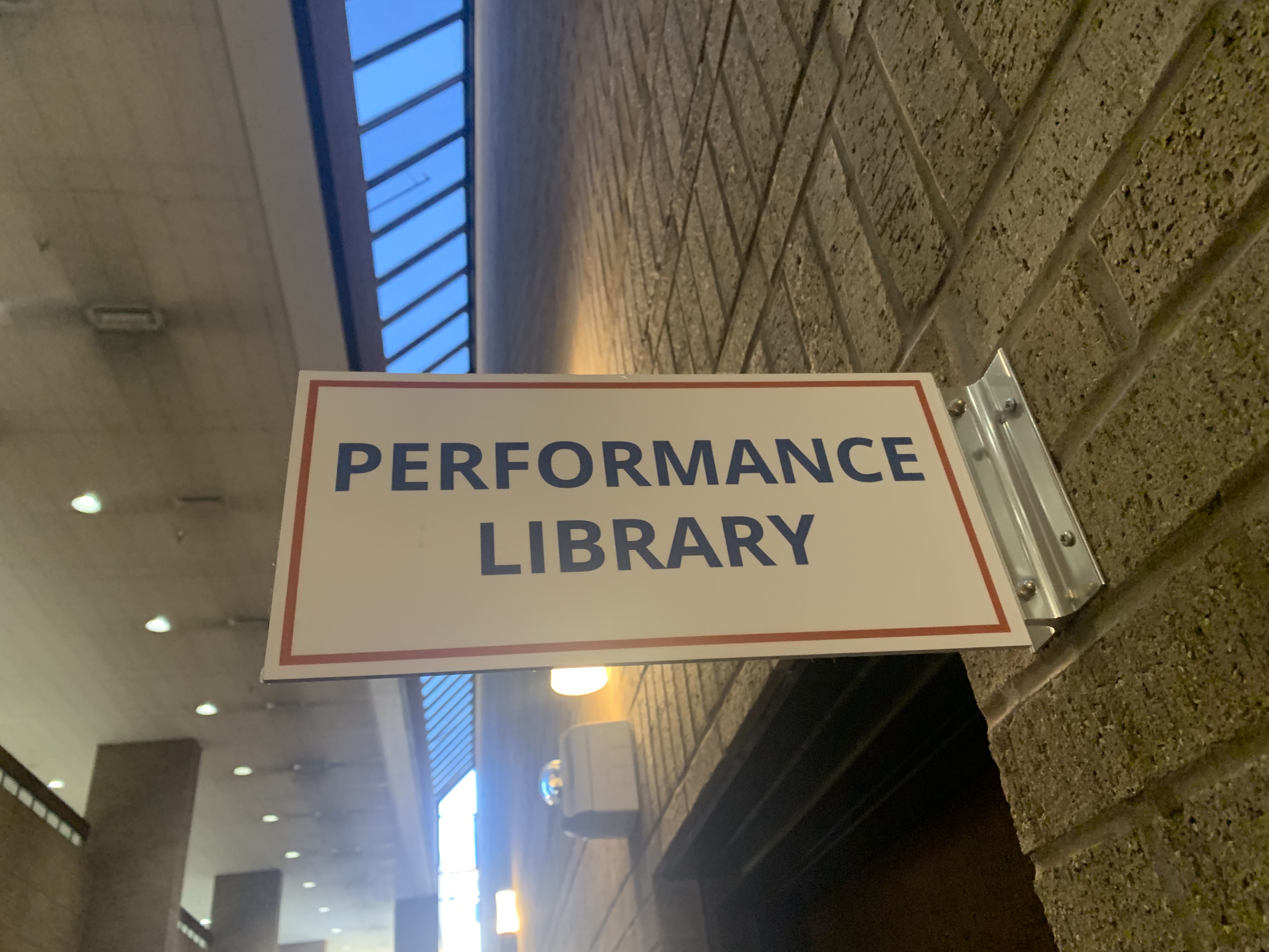 sign outside of a darkened doorway with Helevtica blue font announcing "PERFORMANCE LIBRARY"